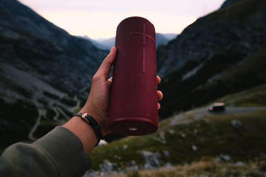 A person holding a red portable speaker with picturesque mountain backdrop.