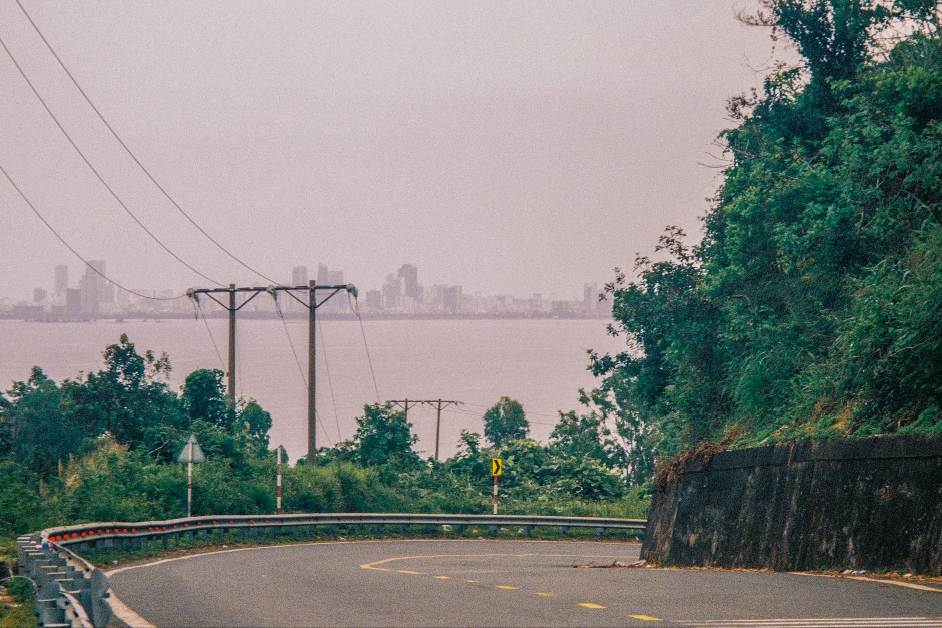 Curvy countryside road with lush greenery overlooking a coastal city skyline under overcast skies.