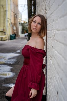 Caucasian woman in a red dress poses against a white wall in an urban alley. Cool and moody setting.