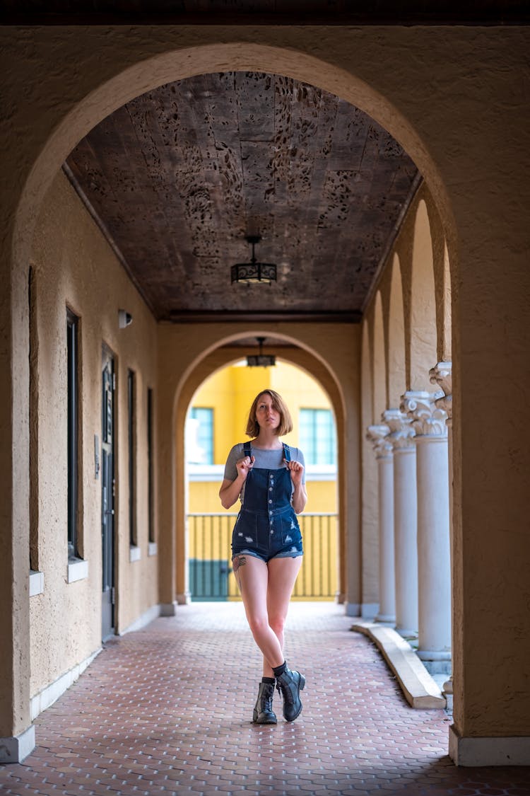 Young Woman In Denim Jumper Shorts Posing On Outside Arched Hallway Of A House 