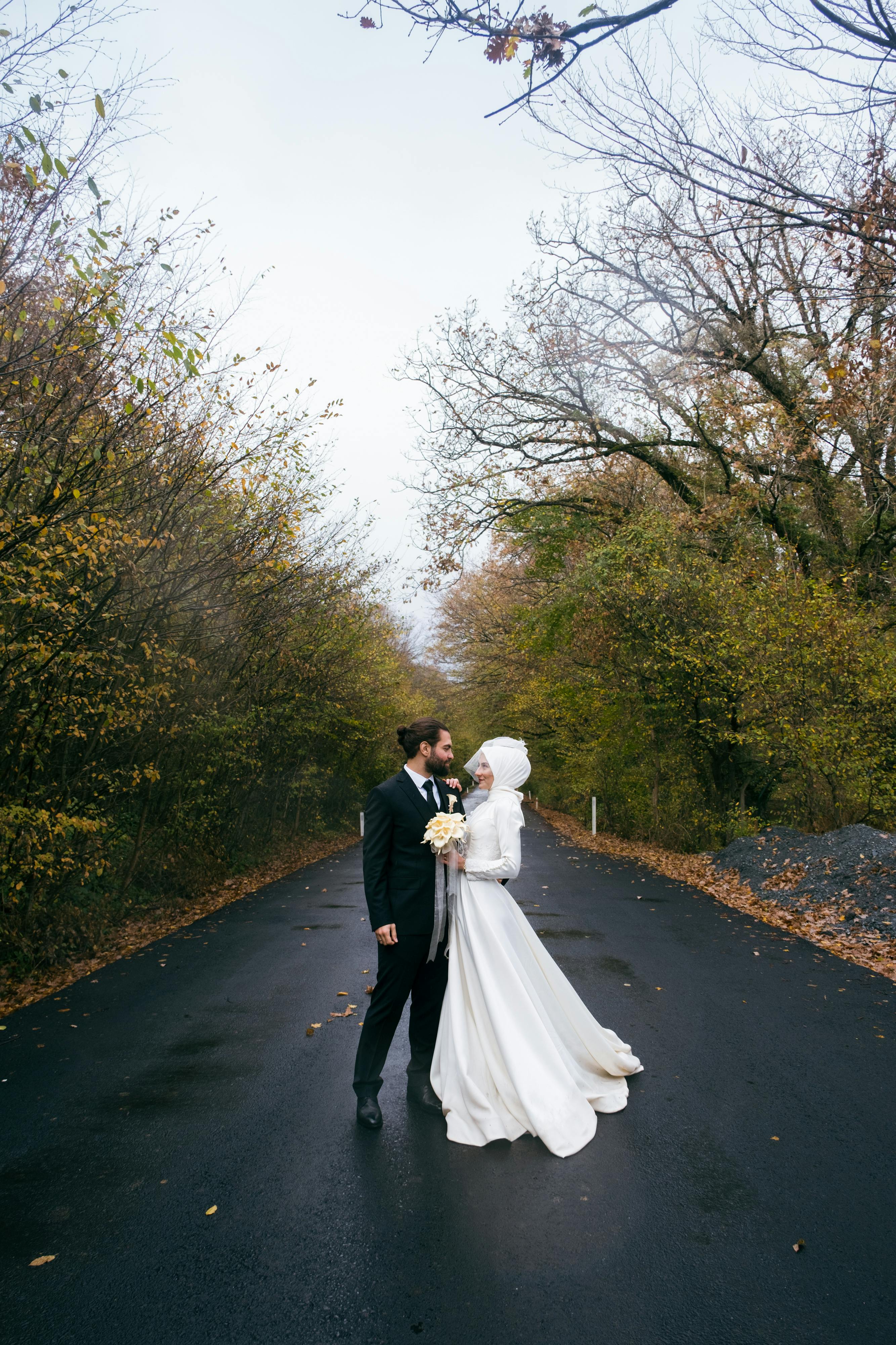 Bride and groom embrace on a tree-lined road in autumn, capturing a romantic moment.