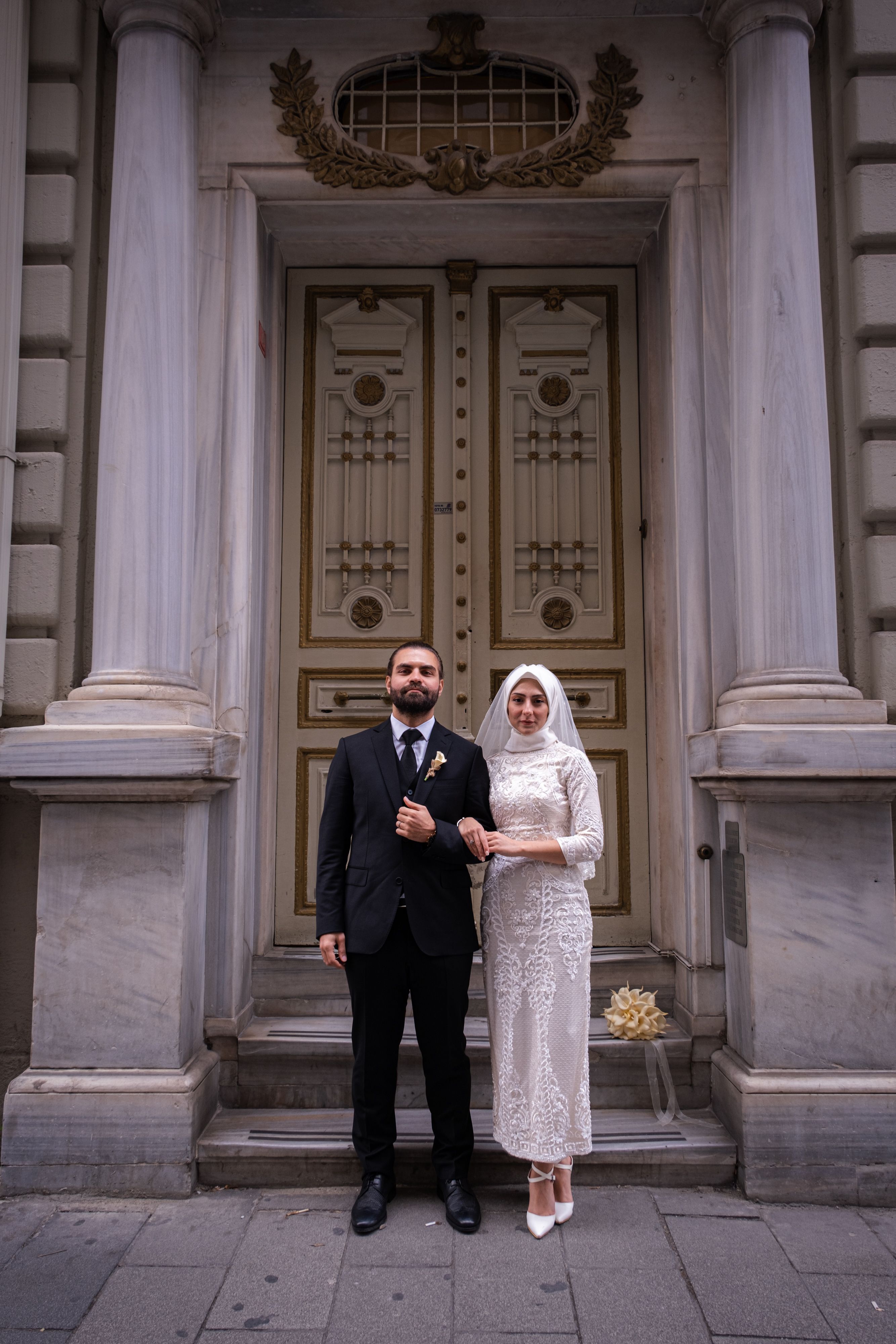 A bride and groom in classic attire pose before a historic doorway, capturing an elegant urban wedding moment.