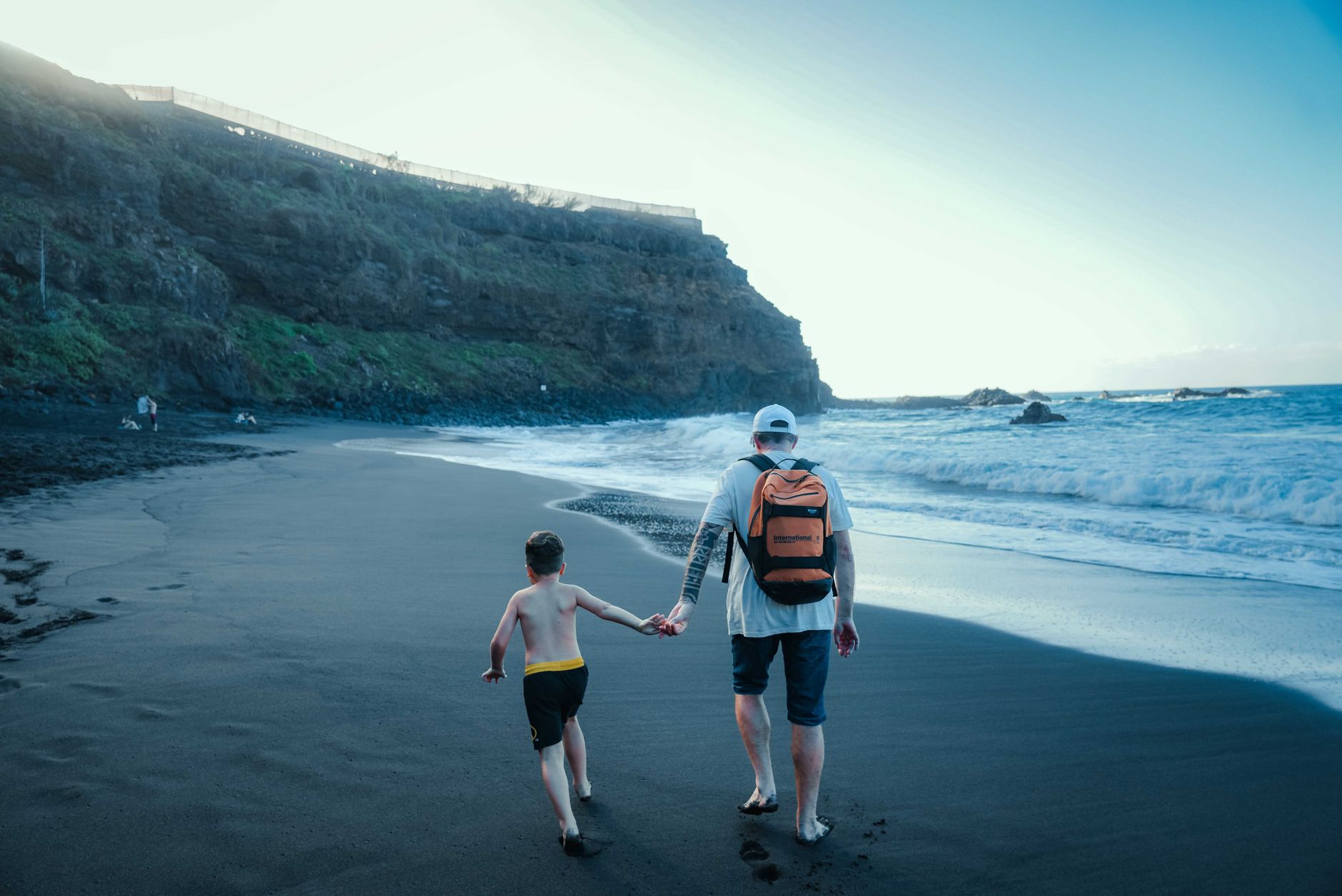 A father and son walk hand in hand on a beautiful black sand beach in Tenerife, Spain.