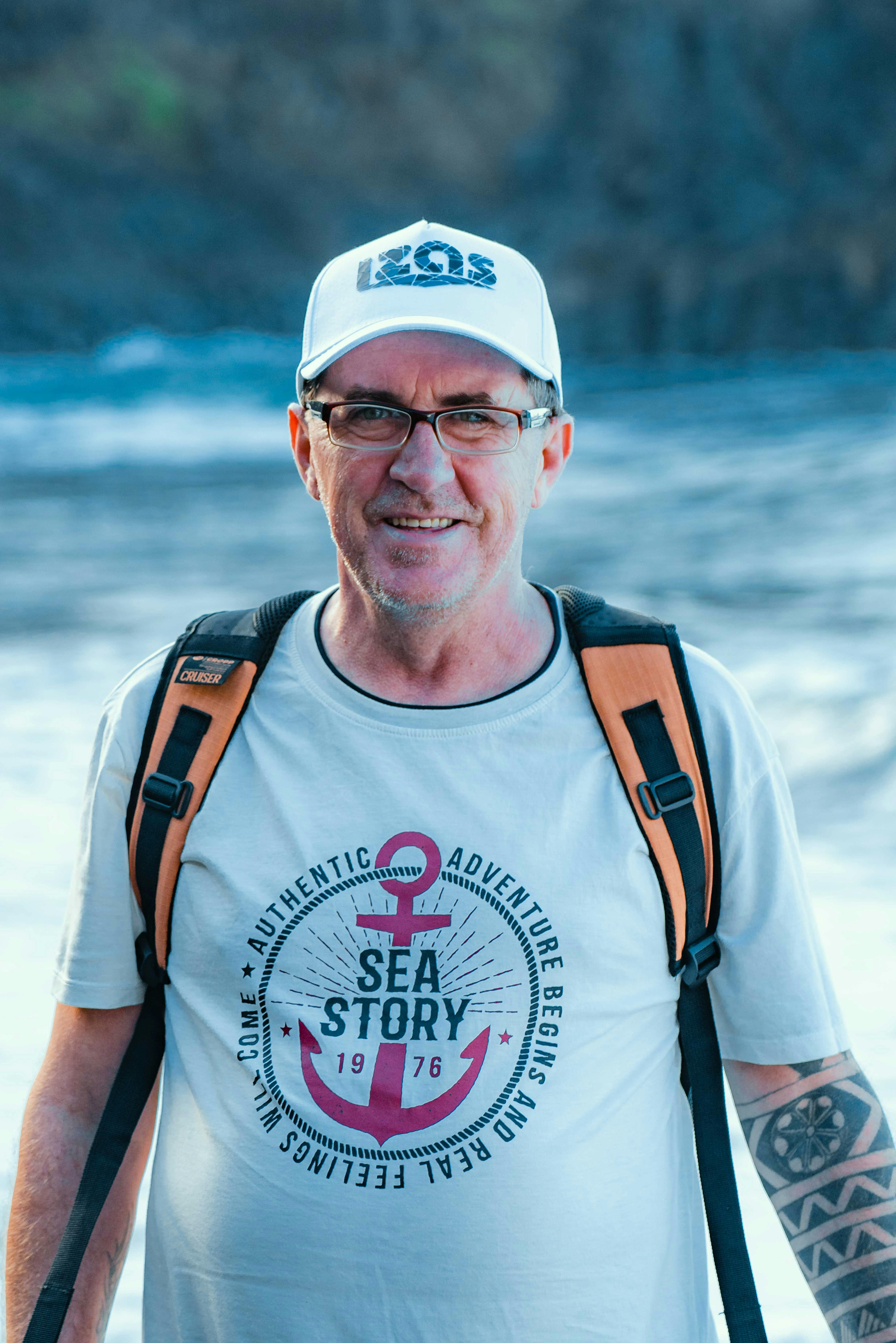 Portrait of a man with glasses, cap, and backpack smiling outdoors near water.