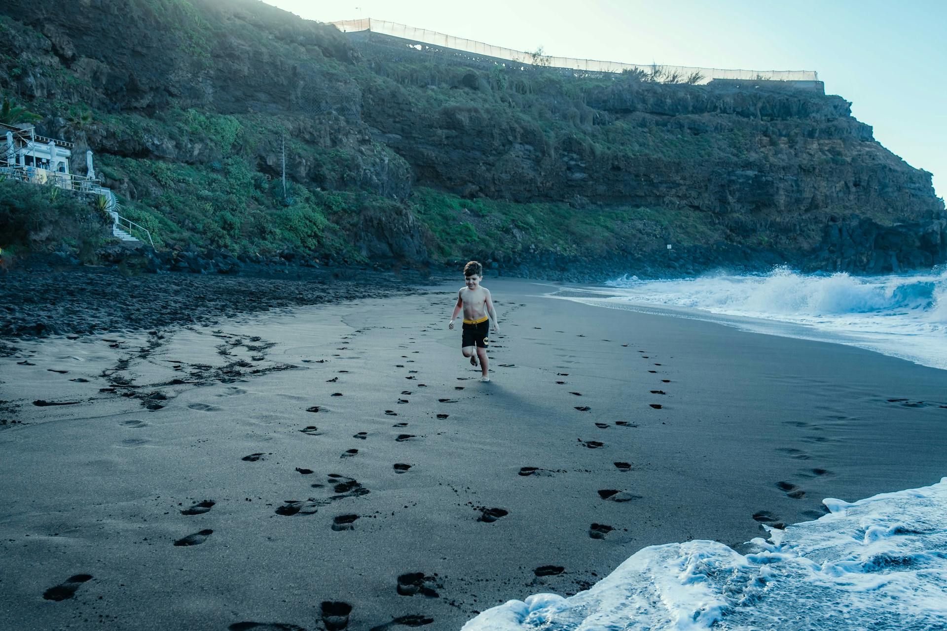 A child joyfully runs along a black sand beach in Tenerife, Spain, leaving footprints behind.