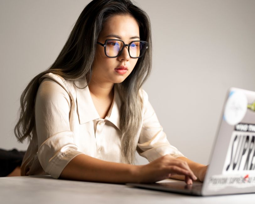 Asian woman wearing glasses intensely working on a laptop at a desk in a modern office setting.