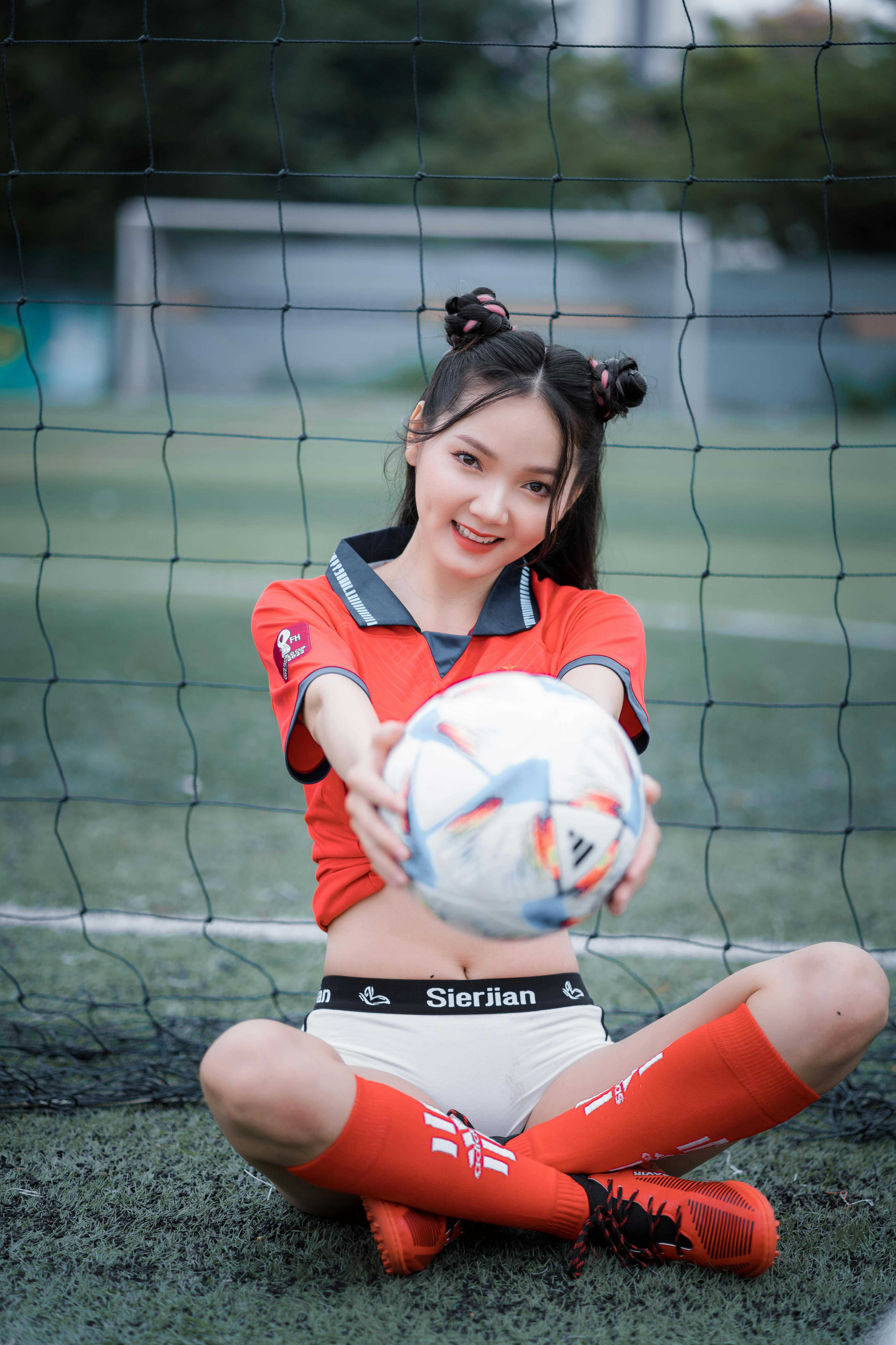 teenage girl in orange jersey shirt and white booty sports short sitting and holding a soccer ball on a field