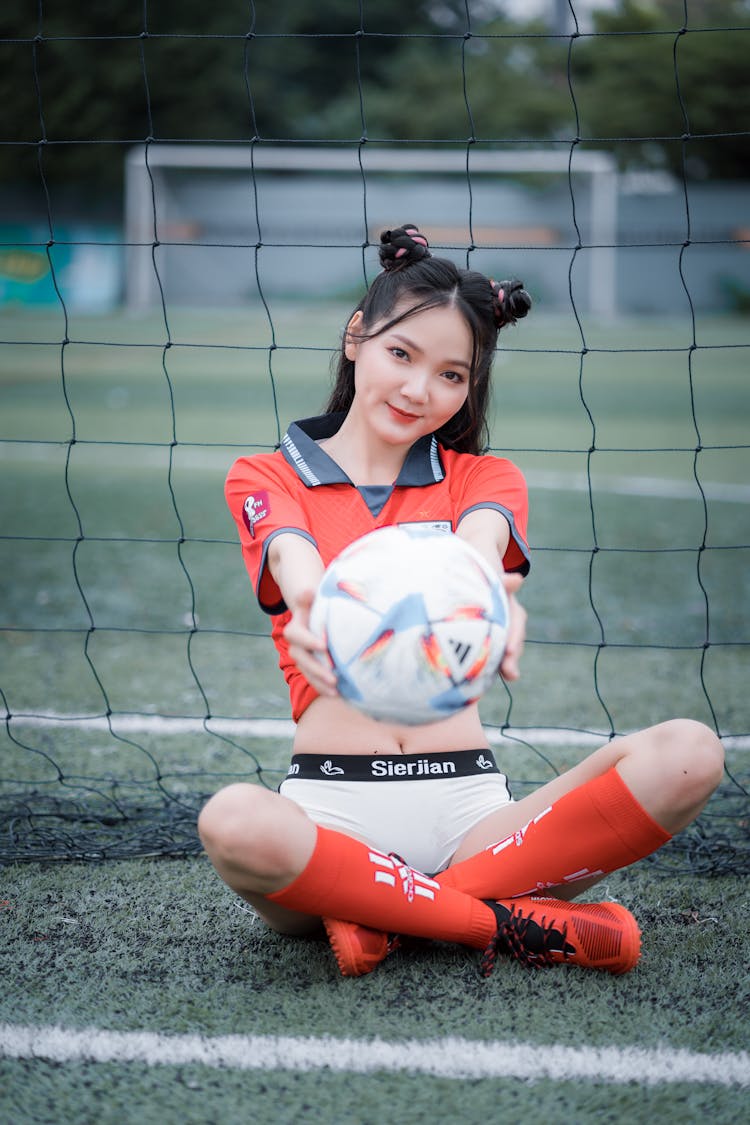 A Woman In A Soccer Jersey Holding A Football While Sitting By A Soccer Goal