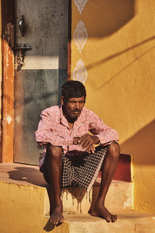 Free Man with black hair in pink shirt sits on sunlit yellow steps beside a wall. Stock Photo