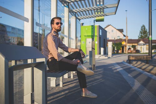 Man Wearing Sunglasses Sitting at Bus Stop during Daytime