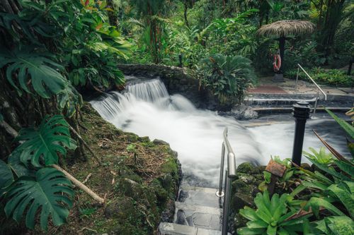 Free Captivating view of a tropical waterfall surrounded by lush greenery, featuring a stone pathway with handrails. Stock Photo