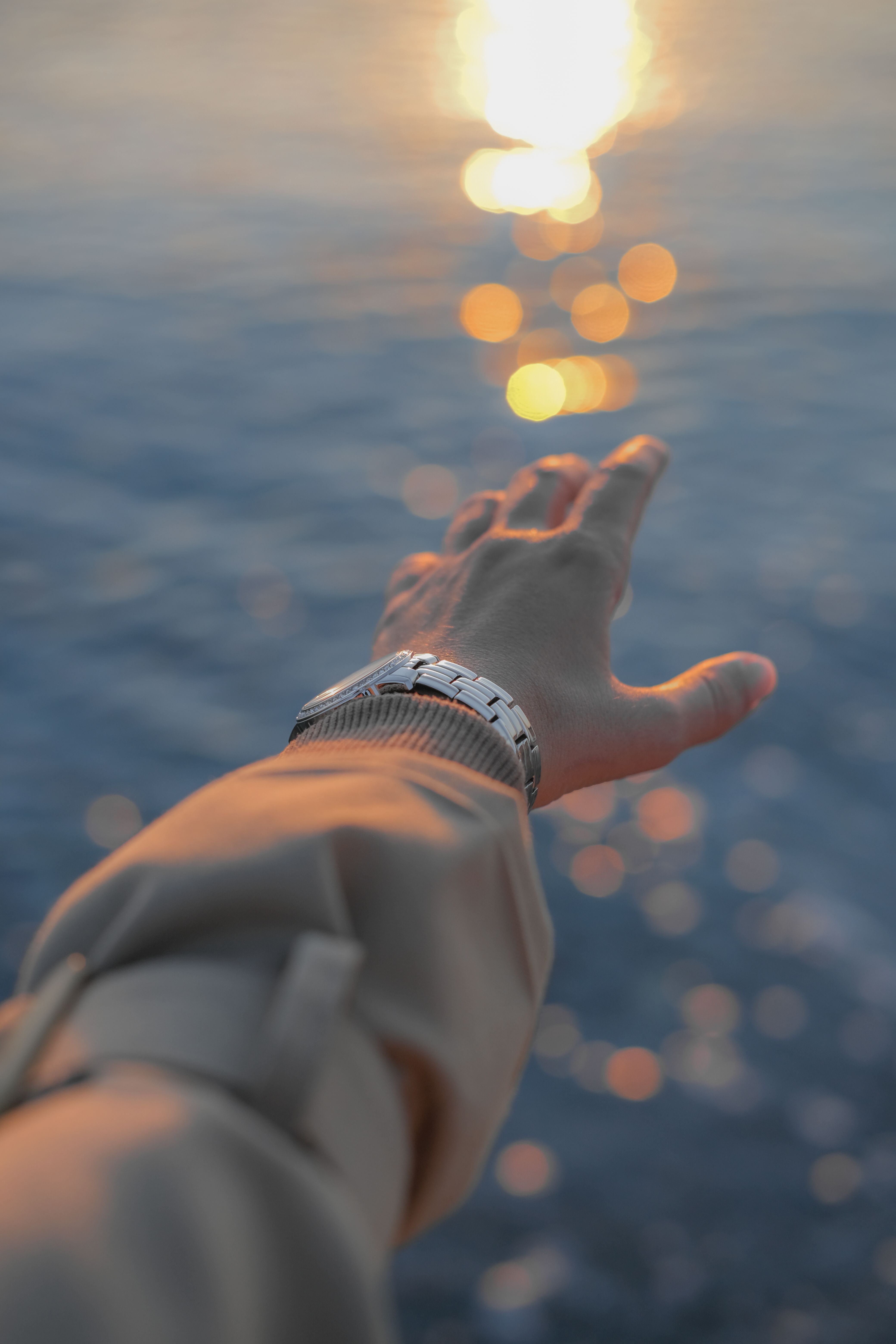 A hand with a wristwatch reaching towards the shimmering water reflecting sunlight.