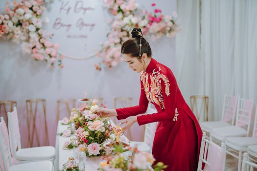 An Asian woman arranges a floral centerpiece for a wedding table wearing traditional attire in a vibrant setting.