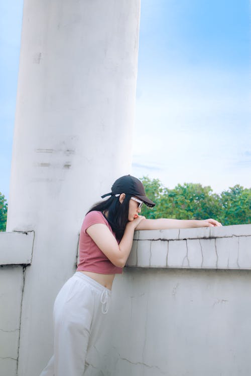 A Woman in Pink Crop Top and Black Cap Leaning on a Wall while Looking Afar