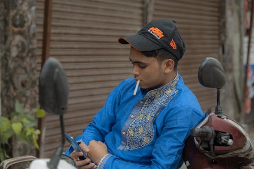 A young man in traditional attire uses his phone while smoking, outdoors on a scooter.
