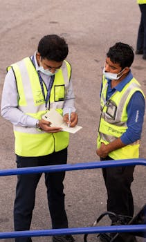 Two airport staff in high-visibility vests reviewing documents while wearing face masks.