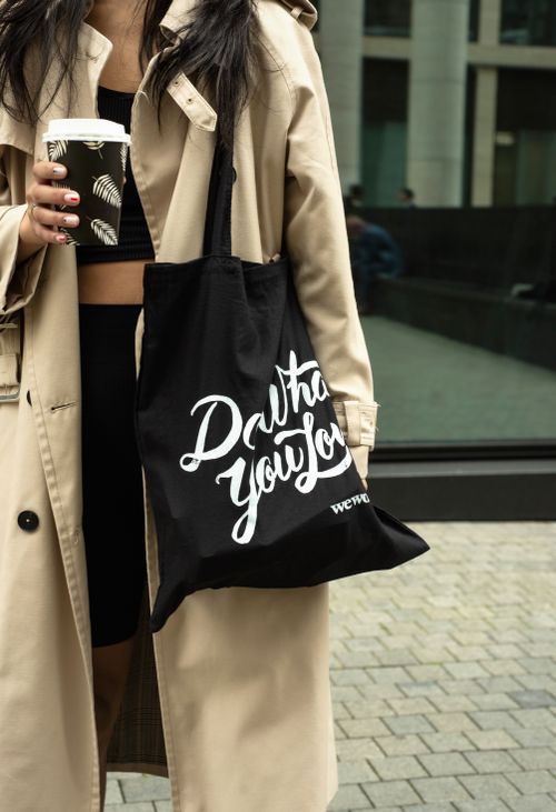 Free Fashionable woman holding a trendy tote bag and coffee cup in an urban setting, wearing a beige coat. Stock Photo