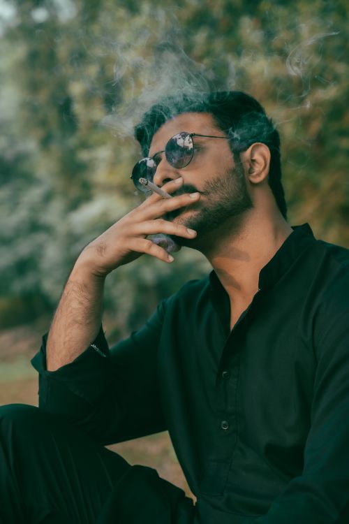 Free Man in black attire and sunglasses nonchalantly smoking outdoors. Stylish and relaxed vibe. Stock Photo