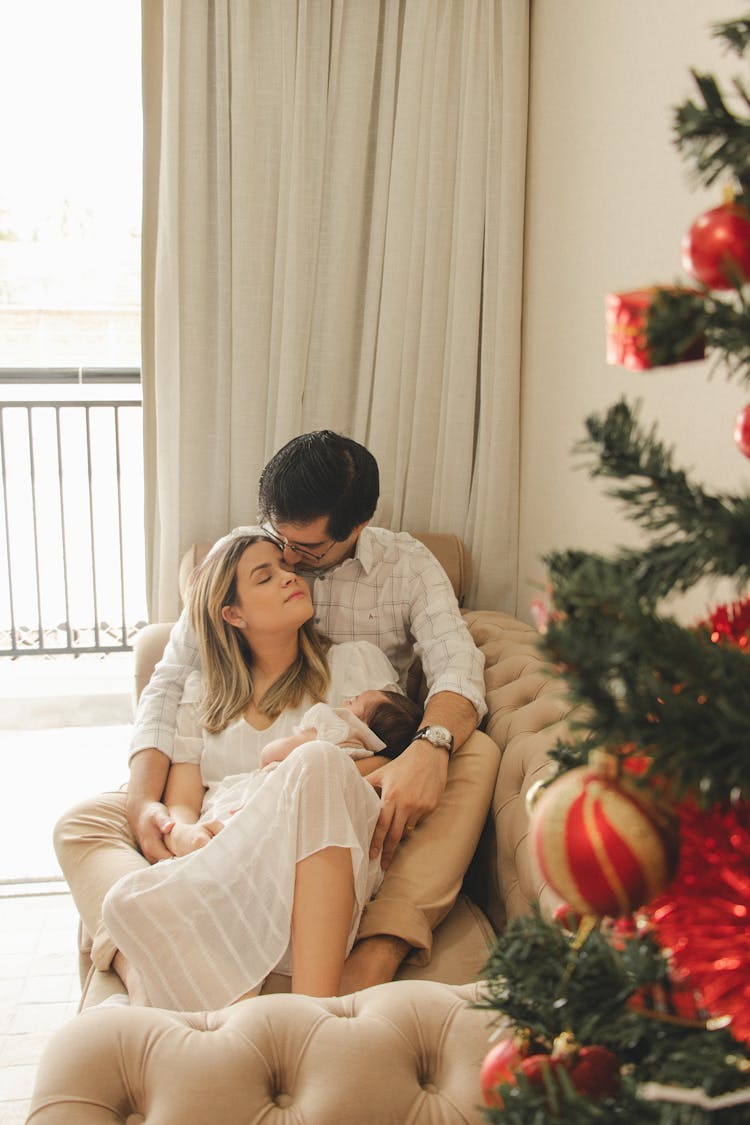 Happy Parents With Newborn On Sofa Near Christmas Tree