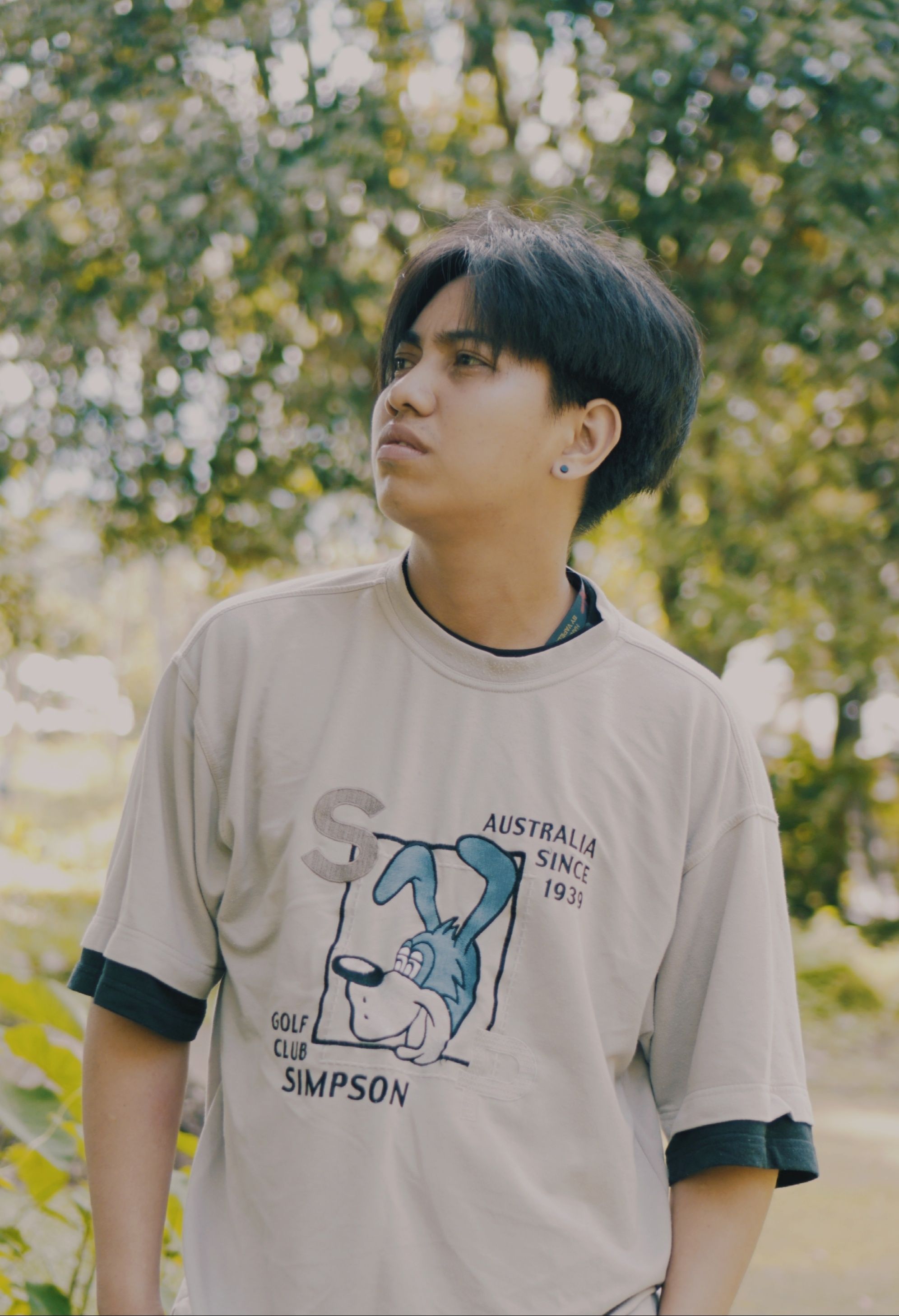 A young man stands outdoors, wearing a graphic t-shirt, looking thoughtfully to the side.