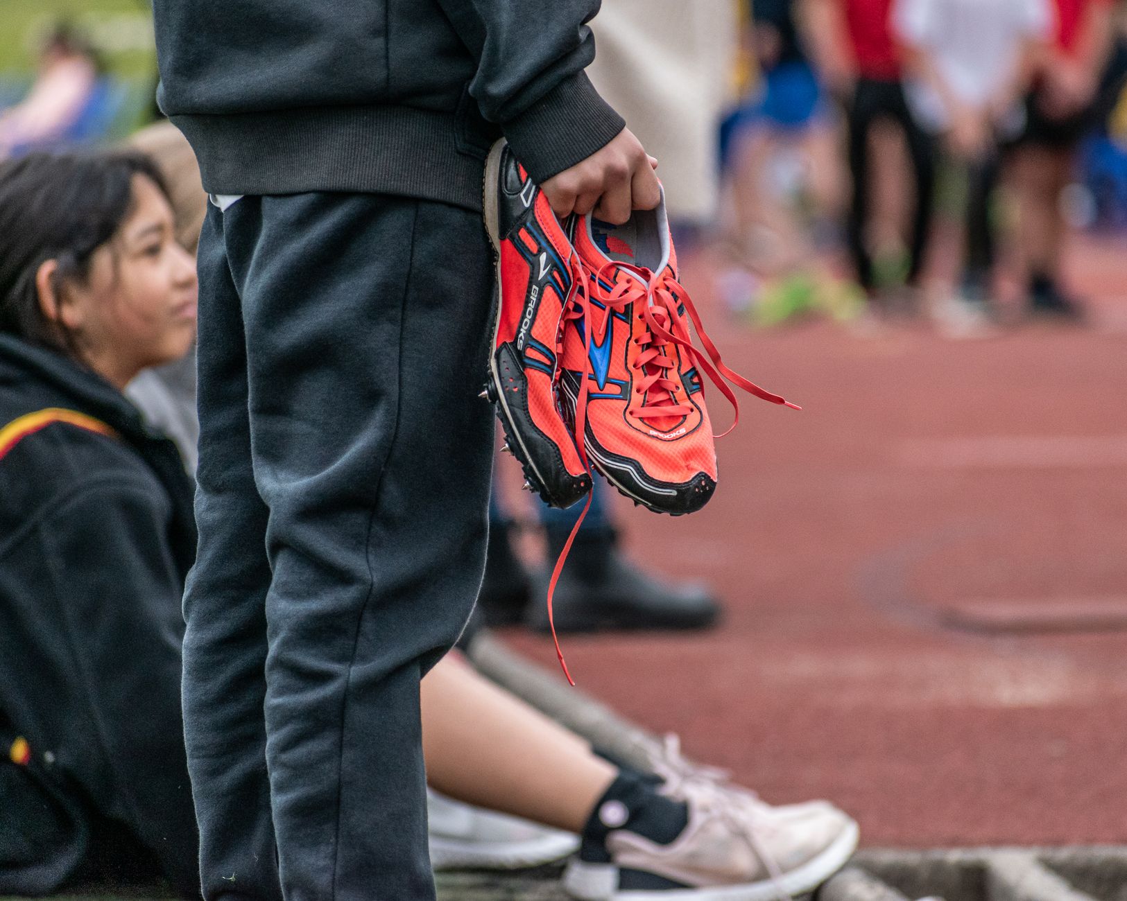 Individual holding red running shoes at a track event with people in the background.
