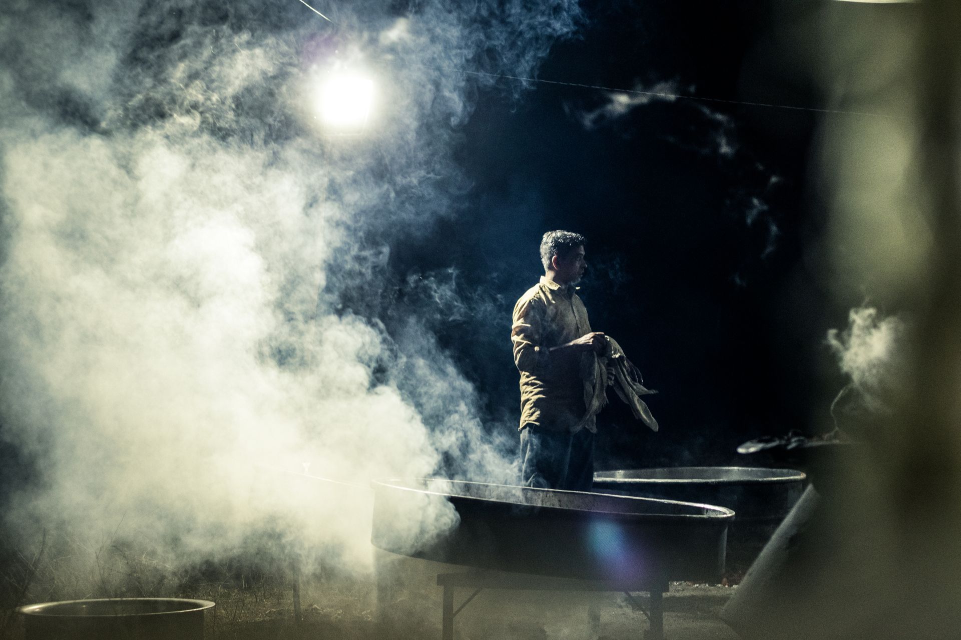 A worker handles fabric amid steam in an atmospheric industrial setting with low lighting.