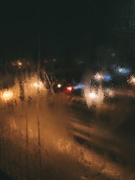 Blurry city lights seen through a wet glass window at night, creating a moody atmosphere.