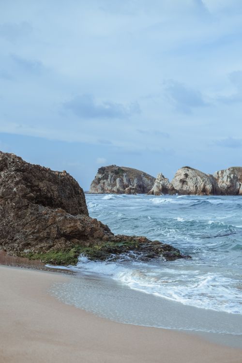 Free Serene view of a rocky beach with ocean waves crashing under a blue sky. Stock Photo