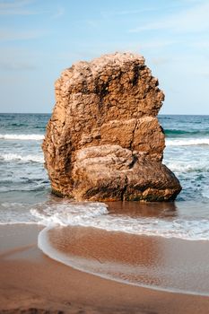 A large rock formation stands prominently on a sandy beach with gentle waves.
