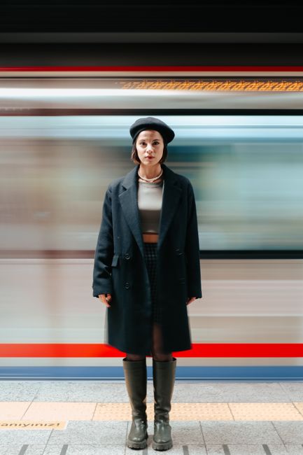 A woman stands in a train station with motion blur background, wearing a black coat and beret.
