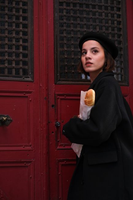 Stylish woman in beret holding baguette near historic red doors, evoking classic urban chic.