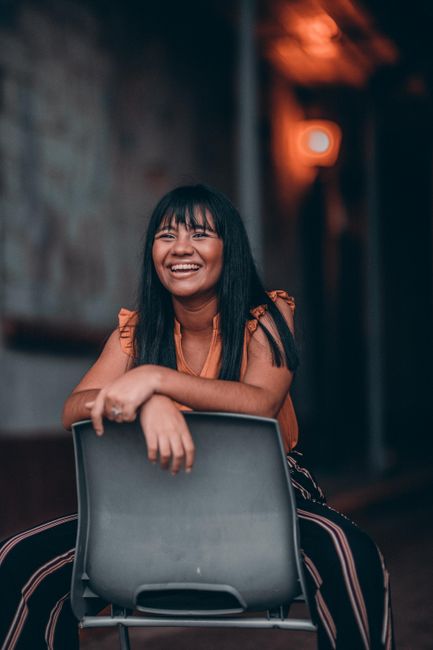 A woman with long hair poses confidently while sitting on a chair indoors, under warm lighting, creating a relaxed, fashionable vibe.