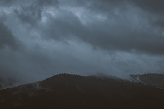 Dark clouds loom over a forested mountain, creating a moody and dramatic scene at dusk.