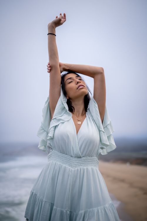 Free Woman in light blue dress with raised arms by the ocean shore, exuding tranquility. Stock Photo