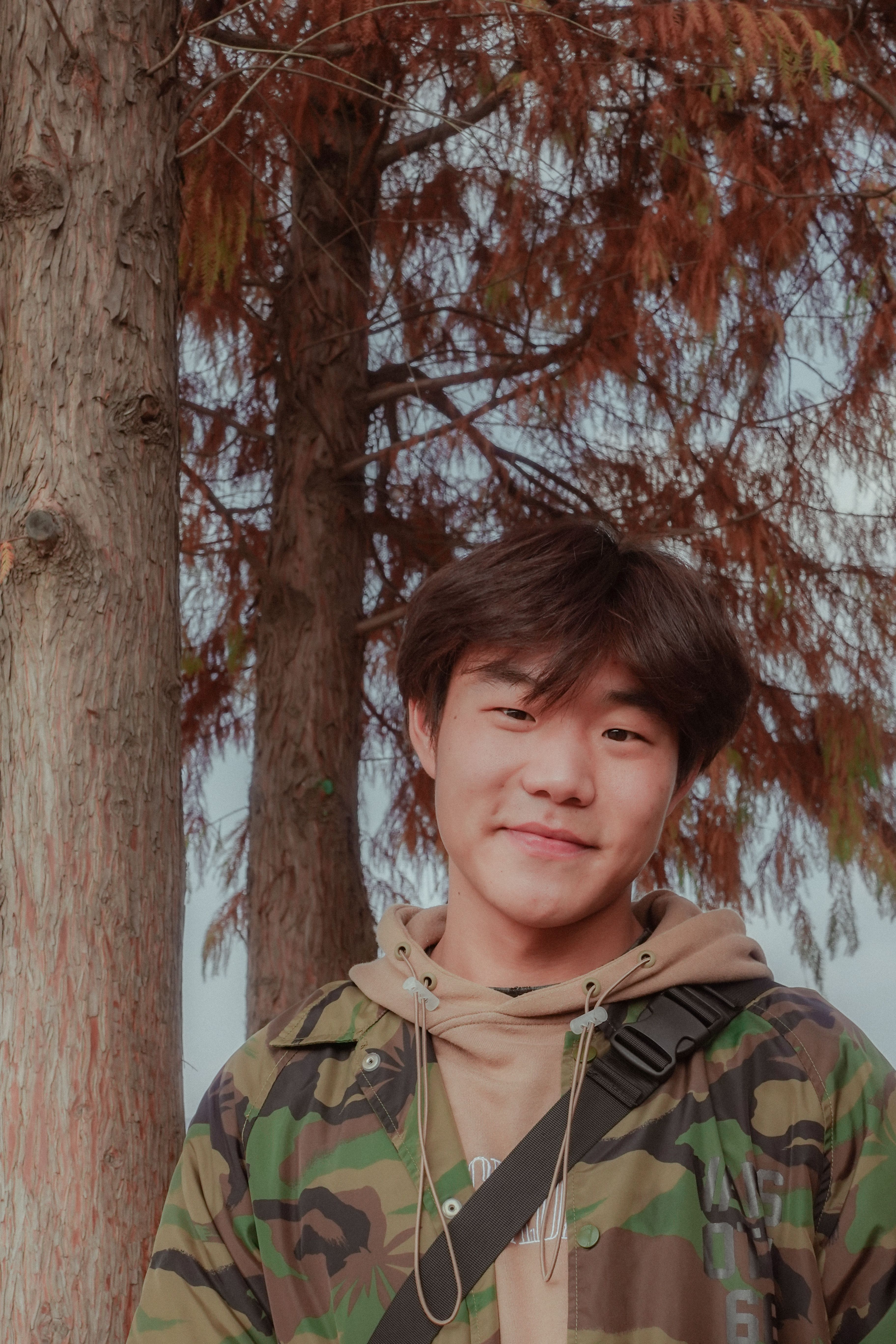 Portrait of a smiling teenager in a camouflage hoodie standing near trees.