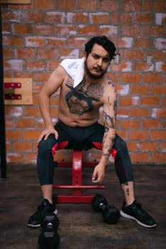 Tattooed man sitting on bench in a gym setting with weights, showcasing strength and fitness.