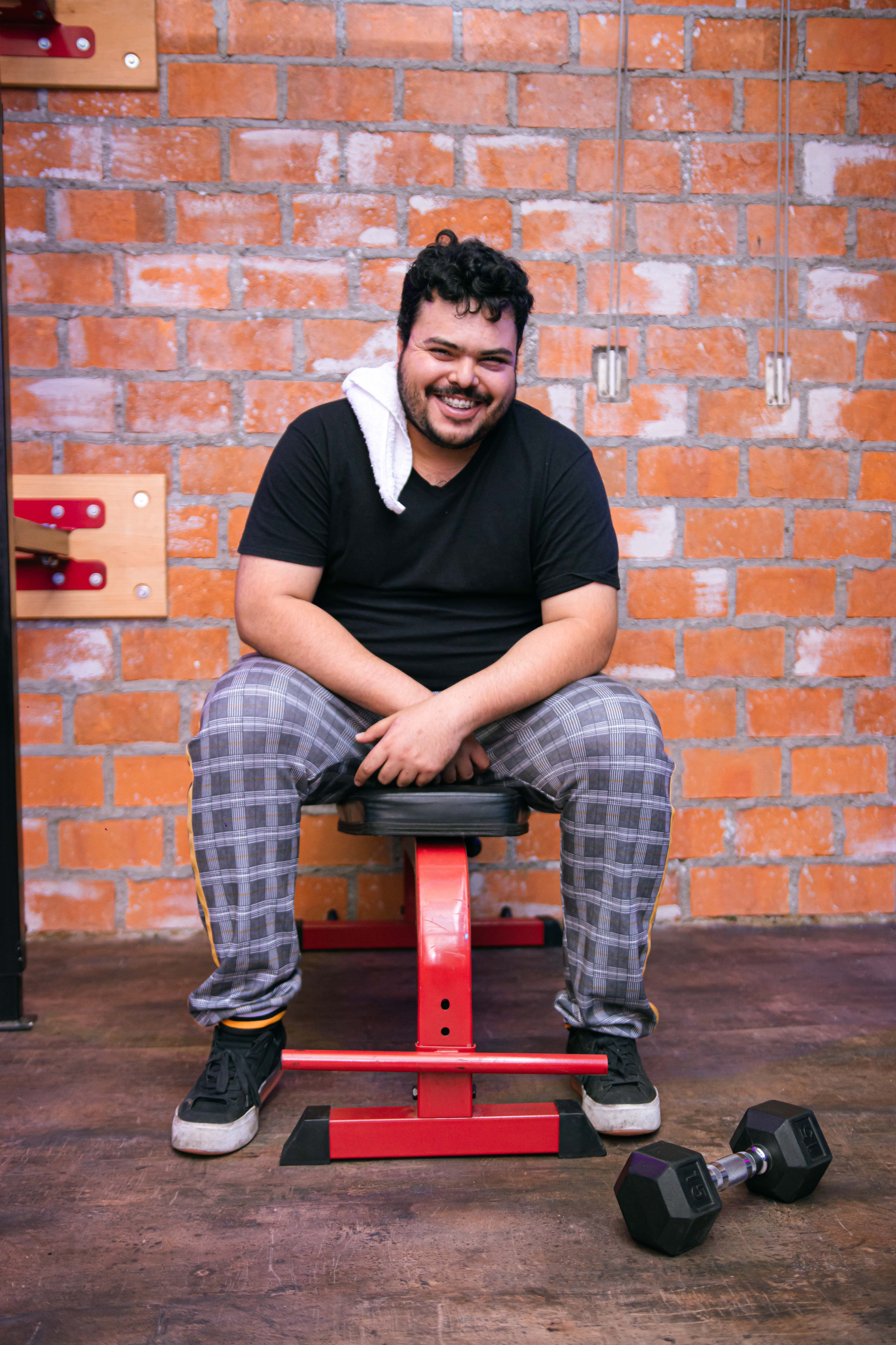 Man with towel and dumbbell, sitting on bench, smiling in a gym setting.