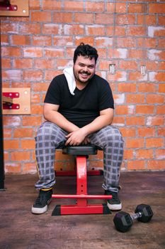 Man with towel and dumbbell, sitting on bench, smiling in a gym setting.