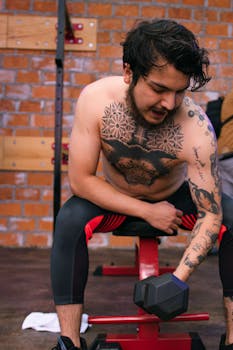 Shirtless man with tattoos lifting a dumbbell indoors. Fitness and strength training focus.