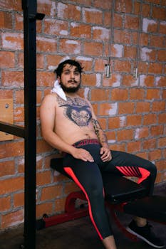 A tattooed man sits shirtless in a gym resting against a brick wall, looking pensive.
