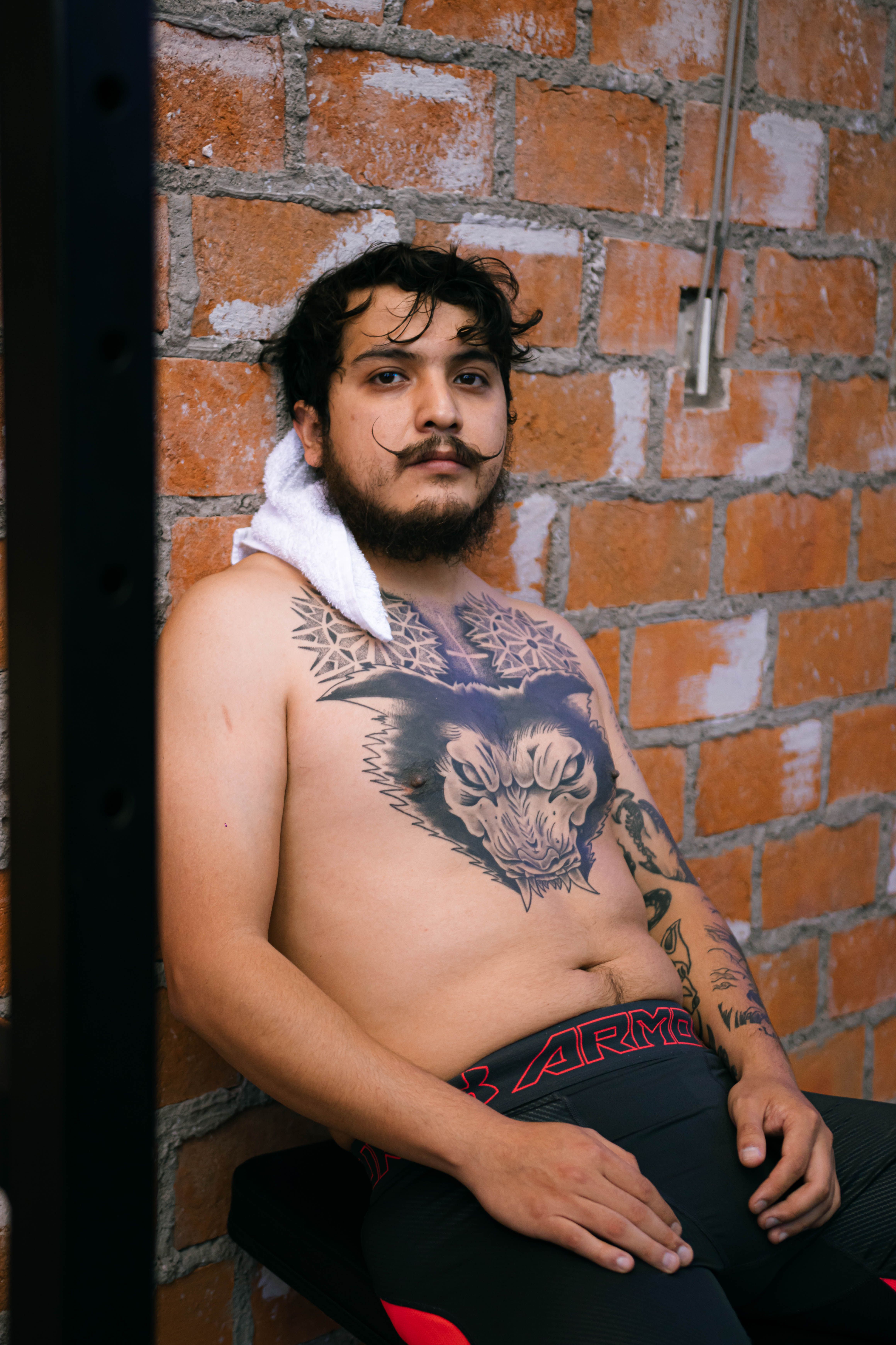 Shirtless man with intricate tattoos sits against a brick wall in a gym setting.