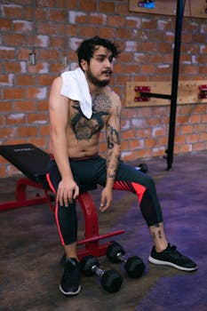 Shirtless man with tattoos resting on bench in gym, looking tired with dumbbells on the floor.