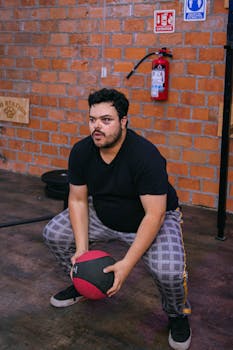 Focused man squatting with a medicine ball in a gym featuring brick walls and fitness equipment.
