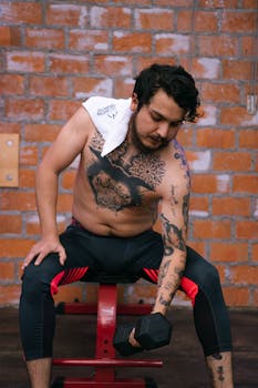 A tattooed man sits shirtless, lifting weights in an indoor gym setting.