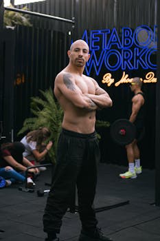 Shirtless muscular man posing indoors at a gym, with others working out in the background.