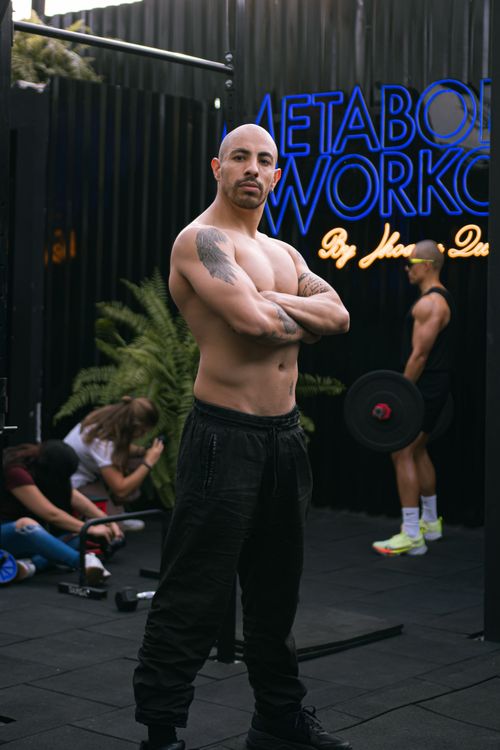 Free Shirtless muscular man posing indoors at a gym, with others working out in the background. Stock Photo