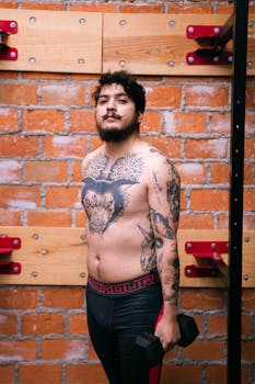 Athletic man with tattoos working out indoors, holding dumbbells against a brick wall.