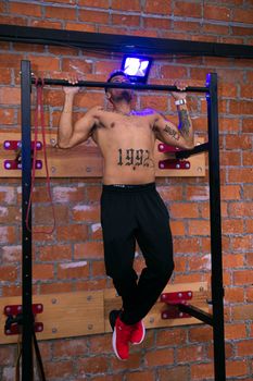 A tattooed man exercising shirtless on a pull-up bar against a brick wall indoors.