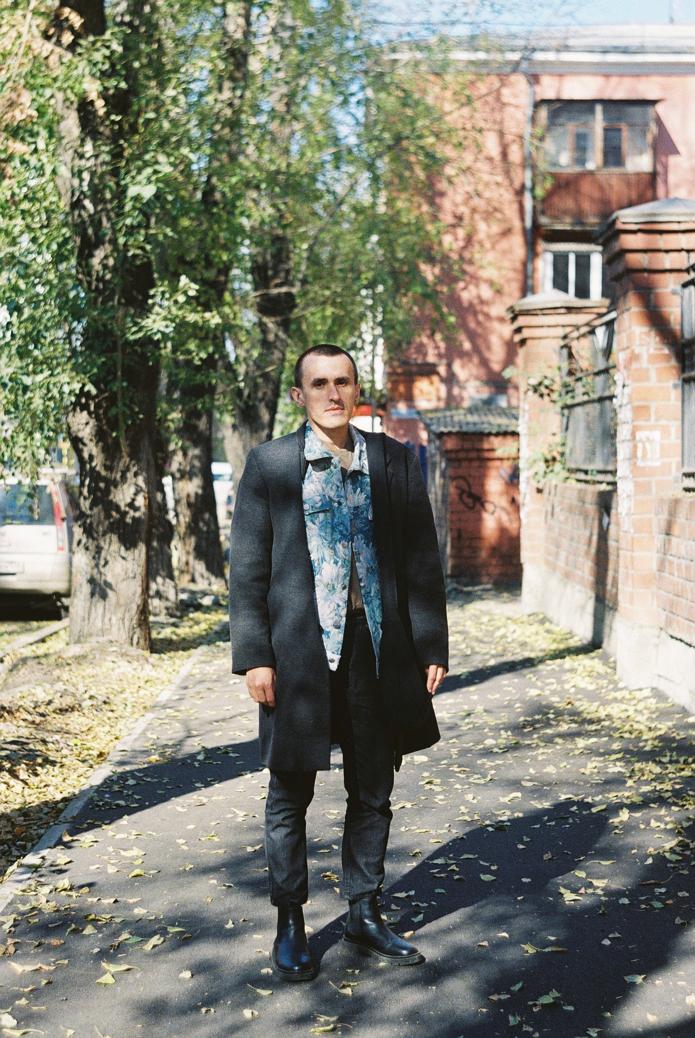 Man in a gray coat stands on an autumn sidewalk surrounded by trees and brick buildings.