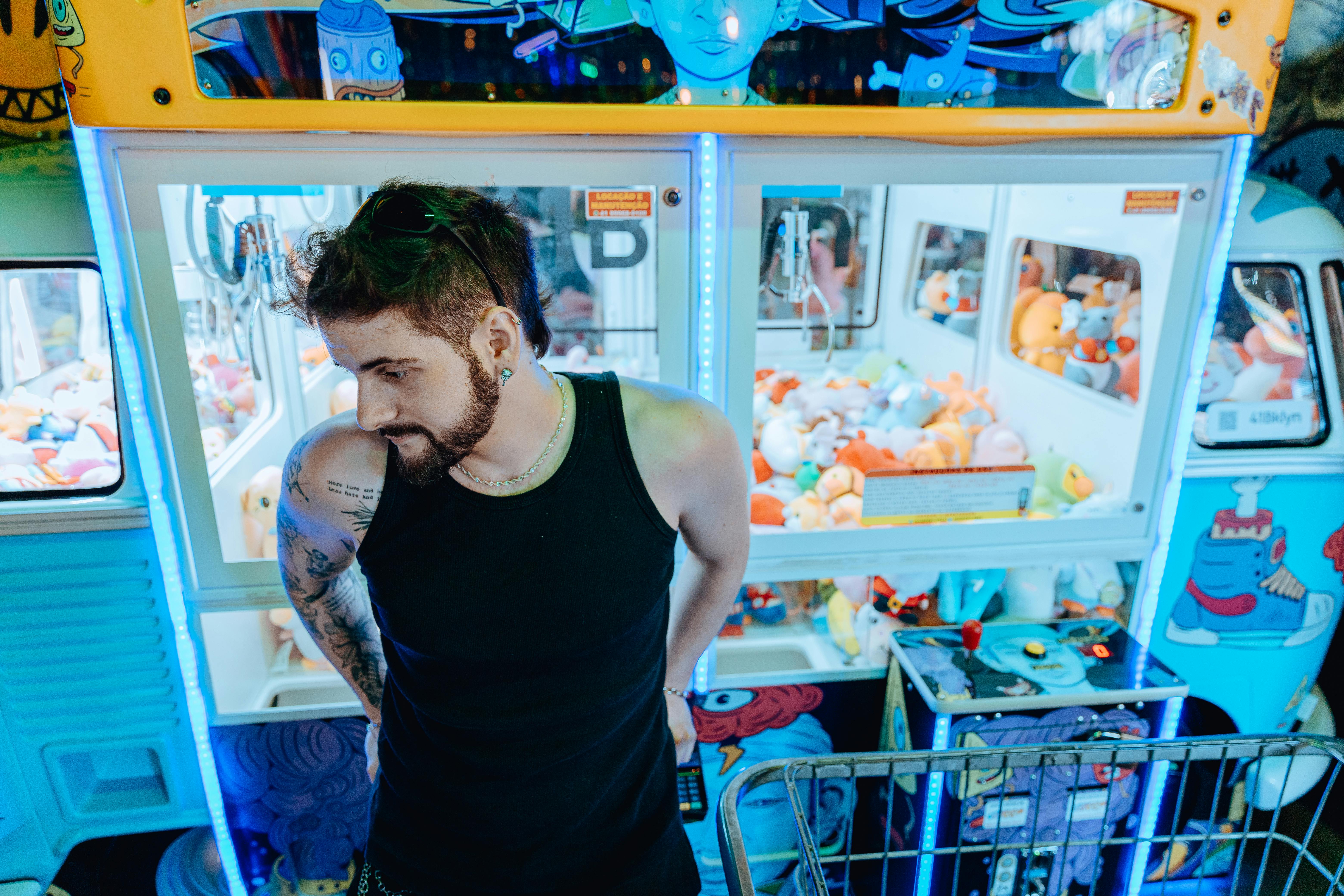 Young man with tattoos standing by colorful claw machine at an amusement park.